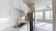This single-floor space showcases a thoughtfully designed interior with a clean, contemporary aesthetic throughout. The kitchen is a standout feature, equipped with stainless steel appliances including a French door refrigerator with ice and water dispenser and a dishwasher. Crisp white shaker-style cabinetry extends to the ceiling, providing generous storage, while granite countertops with subtle veining add a refined touch. A subway tile backsplash in a classic white horizontal layout complements the overall palette, and a stainless steel undermount sink with a modern pull-down faucet completes the space.

Crown molding runs along the ceiling, adding an architectural detail that elevates the unit's finish level. The kitchen transitions seamlessly into an open living area finished with wood-style plank flooring that runs throughout. Large windows dressed with horizontal blinds invite abundant natural light, casting warm sunlight across the neutral-toned walls. The living area offers a sense of openness and airiness, enhanced by the light color scheme and clean lines. HVAC vents are integrated into the ceiling, maintaining a polished and uncluttered appearance across the space.