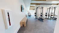 Located at 2500 Bobby Foster Rd SE in Albuquerque, New Mexico, this modern fitness center offers a generously proportioned single-floor layout designed to support a full range of workout activities. The space is divided into three functional zones, each catering to a distinct training focus.

The main cardio area is bright and airy, featuring floor-to-ceiling windows that flood the room with natural light and provide open views of the surrounding outdoor grounds. Treadmills, ellipticals, stair climbers, and rowing machines are arranged to take full advantage of the natural light and views. Rustic exposed wood ceiling beams contrast elegantly with the clean white walls, adding warmth and architectural character throughout.

A dedicated strength training zone houses dual power racks with weight benches and barbells, positioned beneath the striking beam ceiling with pendant lighting. Mirrors and side windows enhance the sense of openness in this section. An adjacent free weights area is equipped with a full dumbbell rack, adjustable benches, a cable/functional trainer machine, and a wall-mounted television, making it ideal for a variety of resistance training exercises.

The durable rubber-speckle flooring throughout provides safety and comfort underfoot. The facility is well-lit with a combination of recessed and linear lighting, and glass partition doors connect the zones seamlessly, ensuring an open and cohesive workout environment.
