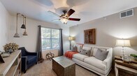 This single-floor residence offers a thoughtfully laid-out living environment with a warm, cohesive aesthetic throughout. The living area is a welcoming centerpiece, featuring plush carpeting, a ceiling fan with light fixture, and a generously sized window dressed with stylish curtains that invite abundant natural light and views of outdoor greenery. The space accommodates both relaxed lounging and flexible use, with a secondary area that functions well as a sitting room or den, also carpeted and naturally lit.

The kitchen is efficiently designed with light wood and white cabinetry providing ample storage, white appliances including a range and dishwasher, a double sink, and wood-look flooring that carries a clean, practical feel. A peninsula counter adds functional prep space.

The bedroom is a comfortable retreat, featuring carpet flooring, a ceiling fan, and a window that continues the natural light theme throughout the home. Generous wall space accommodates storage furniture with ease.

The bathroom is finished with wood-look flooring, a wide vanity with under-sink cabinetry, a large mirror, and a shower-tub combination. The bright, white palette gives the space a clean and airy feel. Overall, the residence presents a balanced blend of functionality and warmth.