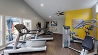 Located in Taylors, South Carolina, this dedicated fitness center is thoughtfully designed to support a well-rounded workout experience. The space is defined by soaring vaulted ceilings that create an open, airy atmosphere, complemented by a striking bold yellow accent wall that energizes the interior. Dark hardwood-style flooring runs throughout, providing a sleek, durable surface that grounds the room's modern aesthetic.

Natural light pours in through a generous arrangement of large windows and a clerestory window positioned high on the wall, offering views of the adjacent outdoor pool area and creating a bright, inviting environment. A ceiling fan ensures comfortable air circulation during workouts, while a wall-mounted HVAC unit provides additional climate control.

The fitness center is well-equipped with a comprehensive selection of Life Fitness cardio and strength machines, including treadmills, elliptical trainers, a multi-station cable machine, and a variety of weight benches and free weights. Full-length wall mirrors amplify the sense of space while allowing users to monitor their form. A wall-mounted flat-screen television adds convenience for entertainment during exercise. Decorative artwork on the walls adds a thoughtful finishing touch, balancing function with an uplifting, polished ambiance throughout the space.