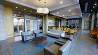This contemporary single-floor space presents a thoughtfully designed lobby and lounge area that blends modern aesthetics with functional comfort. The open-plan layout creates a seamless flow between distinct zones, each serving a unique purpose for residents or guests.

The main lounge area is anchored by a plush gray sofa and coordinating armchairs arranged around a round wood-and-metal coffee table, all set atop a textured blue-toned area rug. Overhead, a trio of elegant drum pendant lights adds a warm, welcoming ambiance to the seating arrangement, complemented by recessed track lighting throughout the ceiling.

Adjacent to the lounge, a dining and social area features bold yellow accent chairs paired with dark tables, introducing a vibrant pop of color against the muted gray and olive-green walls. Pendant lights hang above this zone, reinforcing the cohesive modern design language.

Large glass entry doors at the front of the space allow ample natural light to pour in, enhancing the airy, open feel of the interior. In the background, a bar or service counter area is visible, suggesting additional amenity space for gathering.

The overall ambiance is polished and inviting, with a clean color palette of gray, gold, and olive green that gives the space a contemporary yet approachable character.