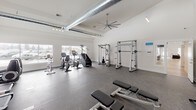 Located at 1681 Pampas Lane in Coeur d'Alene, Idaho, this fitness center is designed to support a full range of workout routines in a bright, open environment. The facility is characterized by dramatically high ceilings that create an airy, spacious atmosphere, enhanced by exposed industrial-style ductwork that adds a modern aesthetic. Large windows along multiple walls flood the space with natural light and offer views of the surrounding area, making workouts feel open and energizing. The flooring throughout is a durable rubberized surface well-suited for heavy equipment and high-traffic use. The cardio zone is generously equipped with treadmills, elliptical trainers, upright and recumbent stationary bikes, and a stair climber, all positioned to take advantage of the natural light from the surrounding windows. The strength training area includes a multi-functional cable machine, a Smith machine, weight benches, and a dumbbell rack, providing comprehensive options for resistance training. Large ceiling fans ensure effective air circulation throughout the facility. A dedicated alcove houses dual drinking fountains for convenient hydration, and a restroom is accessible directly from the main floor. Pendant and track lighting fixtures provide ample illumination throughout, rounding out a well-appointed, functional fitness facility.
