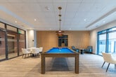 Located at 375 South Street in London, Ontario, this expansive single-floor amenity space is thoughtfully designed to offer residents a refined and versatile communal experience. The layout flows seamlessly across several distinct zones, each with its own character and purpose.

The welcoming lobby/lounge area sets an elegant tone with a coffered ceiling, a sculptural gold ring chandelier, warm wood panel accents, built-in display shelving, and plush mustard-hued armchairs arranged around a central coffee table. Adjacent to the lobby, a spacious open lounge provides generous seating with a mix of sofas and accent chairs in neutral tones, all set atop light hardwood flooring that runs throughout the space.

The games room is a standout feature, centered around a vivid blue billiards table illuminated by a trio of pendant lights. Teal accent walls add a bold pop of color, while large floor-to-ceiling windows flood the room with natural light and offer views of the outdoor area beyond.

The "Oasis" social lounge and dining area is lined with sleek white dining tables and upholstered chairs, complemented by warm wood slat columns. A dramatic black marble-accented kitchen bar with counter seating anchors the back of the room, complete with pendant lighting and full cabinetry — ideal for social gatherings and events. Throughout, the design balances contemporary sophistication with a welcoming, community-oriented atmosphere.