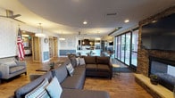 This single-floor communal space is thoughtfully designed to accommodate a variety of social and functional activities. The layout flows seamlessly across several distinct zones, each with its own character while maintaining a cohesive aesthetic throughout.

The main lounge area features a large sectional sofa and additional seating arranged around a stone-clad fireplace wall, complete with a wall-mounted television. Large floor-to-ceiling windows and sliding glass doors flood the space with natural light and provide views of the surrounding exterior. The open-plan design connects this lounge to a central seating area with round tables and upholstered chairs, ideal for casual gatherings.

The fully equipped kitchen is a standout feature, outfitted with dark espresso cabinetry, granite countertops, stainless steel appliances including a commercial-grade refrigerator, a range, dishwasher, and dedicated coffee station. A wall-mounted television adds a functional touch.

A separate dining lounge area offers booth-style banquette seating along textured walls, pendant lighting, and intimate round tables. Warm hardwood flooring runs throughout the entire space, tying each zone together with a clean, welcoming ambiance suited for both relaxed and social use.