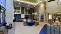 This expansive lobby makes an immediate impression with its dramatic double-height ceilings accented by a tiered, illuminated tray ceiling detail fitted with recessed lighting. The architectural design blends industrial and contemporary elements seamlessly, most notably through the prominent exposed brick columns that anchor the space and add warmth and texture against the smoother, lighter surrounding walls.

A central seating area serves as the focal point of the lobby, featuring a bold navy sectional sofa adorned with decorative pillows in green and earth tones, complemented by a mix of upholstered ottomans and accent cubes in tan and charcoal. This arrangement creates an inviting lounge setting ideal for gathering or relaxing.

The upper level, visible through large steel-framed windows, overlooks the lobby below, adding depth and visual interest to the vertical space. Decorative wall art and a stylized typographic installation on the brick wall contribute to the modern aesthetic. The polished light-toned flooring reflects the ambient lighting, enhancing the open, airy feel of the space. Overall, the lobby exudes a sophisticated, upscale ambiance with a welcoming character.