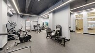 Located at 265 College St in New Haven, Connecticut, this fitness center offers a well-appointed exercise space designed for a comprehensive workout experience. The room features a modern industrial aesthetic, with an exposed dark ceiling revealing ductwork and structural elements overhead, contrasted by crisp white walls and structural columns that add architectural character throughout the floor.

Geometric linear LED lighting runs across the ceiling, providing bright, even illumination throughout the space. Speckled rubber flooring in a neutral tone covers the entire area, offering both durability and safety underfoot. Natural light filters in through windows along the perimeter, connecting the interior to the streetscape outside.

The exercise room is thoughtfully organized into distinct functional zones. A cardio area is equipped with multiple treadmills and stationary bikes, including Echelon spin bikes, all fitted with individual screens. A dedicated strength and resistance training section features machines such as a leg extension/curl unit and additional cable-based equipment. Stability balls in multiple sizes are neatly displayed on a wall-mounted rack alongside mirrors, supporting flexibility and functional training. A flat workout bench rounds out the free-weight area. The space also includes wall-mounted sanitizing stations, reinforcing a clean and well-maintained environment suitable for daily fitness use.