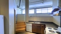 This single-floor space showcases a distinctive industrial aesthetic, characterized by exposed spiral ductwork running along the ceiling and a structural concrete column that anchors the open layout. The kitchenette area is equipped with dark wood cabinetry, a double-basin sink with a modern faucet, and a white dishwasher, offering practical functionality within the raw setting. A row of large, industrial-style windows lines the upper portion of the wall, flooding the space with abundant natural light while reinforcing the loft-like atmosphere. The flooring transitions between materials, with wooden stairs featuring natural wood treads leading to an elevated platform or mezzanine level, adding vertical dimension to the space. The white-painted walls and ceiling contrast with the darker cabinetry and exposed mechanical systems, creating a balanced visual dynamic. The open, unfinished aesthetic of the ceiling and structural elements gives the space a versatile character, suitable for a variety of uses. Overall, the combination of industrial details and practical amenities makes this a functional and visually distinctive environment.