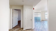 This single-floor, one-bedroom, one-bathroom unit offers a thoughtfully arranged layout comprising a living room, bedroom, kitchen, hallway, and bathroom. The living room is bright and welcoming, featuring wood-style plank flooring, a ceiling fan with light fixture, a large sliding glass door fitted with vertical blinds, and a front-facing window that invites abundant natural light. A wall sconce adds a subtle accent to the open feel of the space. The kitchen is equipped with warm honey-toned wood cabinetry spanning both upper and lower sections, light-toned countertops, a double-basin stainless steel sink, and a dishwasher. A window above the sink provides additional natural light and ventilation. The bedroom is carpeted in a neutral tone with a ceiling fan overhead and a sliding glass door also fitted with vertical blinds offering exterior access and light. A walk-in closet with wood-style flooring and built-in hanging rods provides ample storage. The hallway connects the spaces efficiently with matching wood-style flooring and six-panel interior doors throughout. The unit presents a clean, neutral palette with white walls and trim, offering a functional and comfortable living environment.