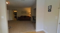 This single-story space features an open-concept layout that connects the living area to the kitchen, creating a seamless flow throughout the interior. The living room is finished with plush wall-to-wall carpeting in a neutral beige tone, contributing to a warm and comfortable atmosphere. A textured popcorn ceiling adds dimension overhead, while clean white baseboards and walls provide a bright contrast to the darker furnishings.

The living area is anchored by a rich, dark brown sofa accented with decorative pillows, complemented by a patterned accent chair positioned nearby. A circular metallic wall art arrangement serves as a focal point on the far wall, adding a decorative touch to the space. Ambient lighting is provided by a floor lamp, casting a warm glow throughout the room.

Transitioning toward the back of the space, the kitchen area is partially visible, featuring cabinetry and appliances. The entryway area showcases hardwood-style flooring, creating a clear visual distinction between the entry and the carpeted living space. Overall, the space presents a cozy, functional layout with a cohesive warm color palette throughout.
