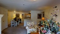 This single-floor residence offers an open-concept layout that seamlessly connects the primary living spaces. The living area serves as the central hub of the home, furnished with a floral-upholstered armchair that anchors the space alongside decorative accents including a table lamp, floral arrangements, and framed wall art, all contributing to a warm, cottage-inspired ambiance. Neutral carpet flooring and beige walls provide a soft, cohesive backdrop throughout.

The kitchen is positioned toward the rear of the open floor plan and features light wood cabinetry extending to the ceiling, a built-in microwave, and a raised counter that functions as a breakfast bar. This counter creates a natural visual divider between the kitchen and the adjoining spaces while maintaining the open feel of the layout.

Adjacent to the kitchen, a compact dining area accommodates a white-painted table and matching chairs, ideal for everyday meals. Warm lighting from flush-mount ceiling fixtures and table lamps enhances the inviting atmosphere. Decorative wall-mounted pieces add character throughout, reinforcing the home's charming and livable aesthetic.