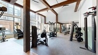 Located at 2902 N Arthur Ashe Blvd in Richmond, Virginia, this fitness center offers a generously proportioned, single-floor workout facility designed with both form and function in mind. The space is defined by its striking architectural character — dramatic exposed dark wood beams cross overhead against soaring vaulted white ceilings, creating a bold visual contrast that elevates the overall ambiance. Ceiling fans are mounted throughout to ensure comfortable airflow during workouts.

Large floor-to-ceiling windows line the perimeter, flooding the interior with abundant natural light and providing open views of the surrounding neighborhood. The flooring transitions between durable rubber fitness flooring in the main workout areas and warm hardwood-style surfaces in transitional zones, providing both safety and aesthetic appeal.

The facility is comprehensively outfitted with a wide array of equipment. Strength training options include Hoist and True-branded cable machines, a multi-functional rack with weight plates, a full dumbbell rack ranging across various weights, and adjustable benches. Cardio equipment includes treadmills, ellipticals, and stationary bikes positioned near the windows to take advantage of natural light and outdoor views. Functional fitness accessories such as kettlebells, resistance bands, stability balls, and additional free weights round out the offerings.

Full-length mirrors are installed along key walls, enhancing the sense of space while supporting proper form during training. The two-tone sage green and white wall treatment adds a calm, modern aesthetic to the environment.