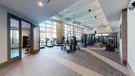 This expansive fitness center offers a comprehensively equipped workout environment across a generous single-floor layout. The main gym area is defined by soaring ceilings with elegant coffered detailing and large-scale floor-to-ceiling windows that flood the space with natural light while providing open views to the surrounding exterior. Ceiling fans are strategically placed throughout to maintain airflow and comfort. The main floor features a wide selection of strength-training machines, free weights, barbells, a full dumbbell rack, benches, a cable system, and a punching bag, all arranged across durable patterned carpet flooring. Large wall mirrors amplify the sense of space and allow for proper form monitoring. A row of treadmills lines the window-facing wall, making cardio sessions bright and engaging. A separate studio room offers a distinct workout environment with warm wood-look flooring, a decorative paneled accent wall, wall-mounted mirrors with a ballet-style barre, spin bikes, a large display screen, and a colorful dumbbell rack — ideal for cycling classes or focused training sessions. A connecting corridor links the two spaces, featuring clean neutral finishes and glass-door entry points, ensuring seamless flow throughout the facility.