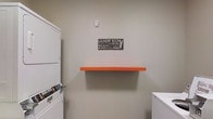 This single-floor laundry room is designed with functionality and practicality at its core. The space features neutral, light gray walls that create a clean and uncluttered atmosphere, making it easy to focus on the task at hand. A stacked washer and dryer unit is positioned to the left, maximizing floor space while accommodating full laundry needs. A striking orange wall-mounted shelf serves as a bold accent piece and provides a convenient surface for laundry essentials such as detergents and fabric softeners. To the right, a white countertop or appliance surface offers additional workspace for folding or sorting laundry. A decorative sign on the wall adds a lighthearted touch, reading "Laundry Room – Loads of Fun: Soak, Wash, Rinse and Dry. Do it yourself. A never ending cycle." The space is well-lit with overhead lighting that ensures good visibility throughout. The overall layout is efficient and straightforward, with every element serving a clear purpose. This laundry room balances utility with a hint of personality, making it a well-appointed and cheerful space for everyday household tasks.