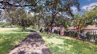 This single-floor property is situated within a beautifully landscaped setting defined by its mature, sprawling trees that provide generous shade over a paved pedestrian pathway. The outdoor environment is a notable highlight, featuring well-maintained green lawns, established hardwood trees with broad canopies, and a classic wrought iron fence with brick pillars that adds a sense of structure and security to the grounds. The pathway winds through the tree coverage, creating a welcoming and walkable exterior atmosphere. Adjacent residential structures are visible beyond the fencing, constructed in warm earth tones that complement the natural surroundings. The overall outdoor ambiance is tranquil and park-like, suggesting a community-oriented setting with an emphasis on green space. The interior of the space could not be clearly captured in the available imagery due to low lighting conditions; however, the property appears to include enclosed interior rooms. The combination of a serene, nature-rich exterior with gated, secured access makes this property a distinctive and inviting space within its neighborhood context.