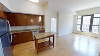 This single-floor space offers a clean, modern aesthetic defined by an open-concept layout that seamlessly connects the kitchen and living areas. The kitchen is well-appointed with rich warm-toned wood cabinetry, granite countertops, a stainless steel dishwasher, and a undermount sink. A central island with a matching granite surface and a lower slatted wood shelf provides additional workspace and storage, anchoring the kitchen as a functional focal point. Pendant lighting hangs above both the island and the adjacent area, adding layered illumination and a contemporary design touch. Light hardwood flooring runs throughout the space, contributing to a bright and cohesive feel. The living area extends generously from the kitchen, offering an open and airy atmosphere with tall ceilings that enhance the sense of volume. A large window along the far wall allows natural light to fill the space, creating a welcoming and well-lit environment. The neutral wall palette keeps the interior feeling fresh and versatile. The overall design balances practicality with modern style, making it a highly functional and visually appealing space suitable for a variety of uses.