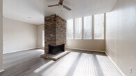 This two-story home offers a thoughtfully designed layout with distinct character across both levels. The main living area immediately draws attention with its soaring vaulted ceiling, anchored by a striking full-height brick fireplace that serves as a bold architectural focal point. A ceiling fan complements the airy volume overhead, while a large bank of floor-to-ceiling windows along one wall floods the space with generous natural light, casting dynamic patterns across the smooth hardwood-style flooring. The open layout of the living area provides a versatile, expansive feel well-suited for both everyday living and entertaining. Adjacent spaces continue the hardwood-style flooring, maintaining a cohesive and modern aesthetic. Moving to the upper level, the bedrooms transition to plush carpeting, creating a warmer, quieter atmosphere. The carpeted rooms are well-proportioned, each featuring ceiling fans and multiple windows fitted with blinds that offer both light control and privacy. A full bathroom with a tub-shower combination is conveniently accessible from the bedroom level. Neutral wall tones throughout the home provide a clean, move-in-ready canvas. A staircase connects the two floors, adding to the functional flow of this well-structured, light-filled home.
