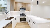 This contemporary three-bedroom, three-bathroom home in Seattle's Lake City neighborhood spans three well-appointed floors, offering a thoughtful layout with an office, dedicated laundry room, and ample closet storage. Light-toned hardwood flooring flows throughout the main living areas, complementing the clean white walls and recessed lighting that give the home a bright, airy feel. The open kitchen is a standout feature, showcasing two-tone cabinetry — crisp white lower cabinets paired with warm wood uppers — white quartz countertops, a subway tile backsplash, stainless steel appliances including a gas range, and pendant lighting over a central island. A half bath on the main level features minimalist matte black fixtures consistent with the home's modern aesthetic. The upper floors host the bedrooms, including a primary suite with plush carpet, generous closet space, and a private ensuite bath featuring dual undermount sinks, white quartz counters, matte black hardware, large framed mirrors, and dark tile flooring. Additional bedrooms offer views of the surrounding greenery through large windows. Each floor maintains a cohesive, modern design language with clean lines and neutral tones throughout.