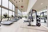 Located at 14020 Stream Way in Charlotte, NC, this state-of-the-art fitness center offers a spacious and thoughtfully designed exercise environment spanning a single open floor. The facility's most striking architectural feature is its dramatic double-height ceiling paired with expansive floor-to-ceiling windows that flood the interior with natural light and provide sweeping views of the surrounding neighborhood. Stylish pendant lighting and ceiling fans complement the bright, airy atmosphere throughout.

Light wood-tone flooring runs the length of the space, lending a clean and contemporary aesthetic. The main exercise area is comprehensively outfitted with a full row of treadmills and elliptical trainers positioned directly in front of the large windows, allowing users to enjoy the outdoor views during cardio sessions. Strength training is well-represented with a multi-station cable machine, free weight benches, and multiple racks of dumbbells.

A dedicated studio room, separated by frosted glass doors, houses spin bikes and a rowing machine alongside a large wall mirror and mounted flat-screen display — ideal for group fitness or individual cycling sessions. The overall ambiance is bright, modern, and energizing, making this fitness center a premium amenity for health-conscious users.