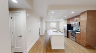 This 790-square-foot, one-bedroom, two-bathroom apartment is located at The Residences at Bella Terra in Huntington Beach, California. The open-concept kitchen and living area serves as the heart of the unit, anchored by a sleek, curved white quartz island with an integrated sink. Warm-toned wood cabinetry, stainless steel appliances — including a gas range, microwave, and refrigerator — and recessed lighting complete the well-appointed kitchen. Wood-style flooring flows throughout the main living areas, adding warmth and continuity. The 160-square-foot primary bedroom features plush carpet, a ceiling fan, and a walk-in closet, while a dedicated en-suite bathroom offers a white quartz vanity, rich wood cabinetry, and ample storage. A second bathroom includes a full soaking tub framed by neutral large-format tile. A private balcony with decorative iron railings overlooks the attractively landscaped courtyard of the Mediterranean-inspired building exterior. The unit benefits from abundant natural light through multiple windows, and the overall aesthetic is clean, neutral, and move-in ready.