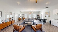 Located at 770 S 40th St in Springdale, Arkansas, this thoughtfully designed single-floor space offers a welcoming, open-concept layout that seamlessly integrates multiple functional zones. The generous living area serves as the heart of the space, featuring soaring vaulted ceilings, warm wood-look flooring, and an abundance of natural light streaming through multiple windows fitted with plantation shutters. Mid-century modern furnishings — including leather accent chairs, a plush gray sofa, and a sleek glass coffee table — lend a sophisticated yet approachable character. A stylish multi-bulb chandelier and ceiling fan complement the airy atmosphere. A dedicated workspace is tucked to one side, complete with a substantial wood desk, guest seating, and its own striking sputnik light fixture, making it ideal for professional use. The kitchen is well-appointed with crisp white cabinetry, granite countertops, stainless steel appliances including a refrigerator, range, microwave, and dishwasher, and a deep sink — offering full functionality in a clean, modern aesthetic. Adjacent to the kitchen, a dining area with a dark-topped table and chairs provides ample seating. A secondary lounge nook with upholstered chairs and a wall-mounted TV rounds out this versatile, multi-purpose space.