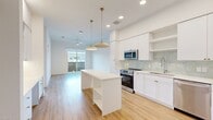 This well-designed 1-bedroom, 1-bathroom unit at Evergreen offers a bright, modern living experience across a single floor. The open-concept kitchen and living area serves as the heart of the home, anchored by a white center island with built-in shelving and illuminated by a pair of stylish brass pendant lights. The kitchen is outfitted with stainless steel appliances, white shaker cabinetry with gold hardware, a tile backsplash, and a deep sink with a professional-style faucet. Light hardwood flooring flows throughout the main living areas, enhancing the airy, clean aesthetic. A ceiling fan and sliding glass doors lead to a private balcony, allowing natural light to fill the living space. The bedroom features plush carpeting, a ceiling fan, and a large window that brings in additional natural light. Two closets provide ample storage throughout the unit. The bathroom is finished with large-format tile flooring, a white vanity with gold hardware, and an illuminated mirror, with a stacked in-unit washer and dryer conveniently nearby. A built-in desk area off the kitchen adds functional workspace, rounding out this thoughtfully appointed home.