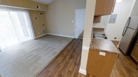 This single-floor space showcases an open-concept design that seamlessly connects the kitchen and living areas. The living area is finished with neutral beige carpet, while the adjoining space features warm wood-look laminate flooring that extends toward the front entry door, creating a clear visual distinction between zones. Walls are painted in a warm tan hue, complemented by clean white baseboards and trim throughout.

The kitchen area is partially separated by a structural column and features wood-toned cabinetry, a stainless steel sink, and granite-style countertops, offering a functional and practical workspace. Multiple floor-level and wall-mounted HVAC vents are positioned throughout, ensuring efficient climate control across the space.

Natural light enters through a window on the left side of the living area, contributing to the overall brightness of the unit. The front entry door is a classic white panel style, providing a clean, understated focal point at the far end of the room. The layout is straightforward and efficient, making good use of the available space throughout.