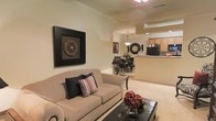 This single-floor residence offers a thoughtfully designed open-concept layout that promotes a natural flow between the living, dining, and kitchen areas. The living room is anchored by a plush, neutral-toned sofa accented with decorative pillows, complemented by a sleek dark ottoman coffee table adorned with floral accents. A bold, framed circular wall art piece adds a striking focal point to the space, reinforcing the sophisticated yet comfortable aesthetic throughout.

Crown molding runs along the ceiling, adding an elegant architectural touch, while soft carpeting underfoot enhances the warmth of the living area. The space transitions seamlessly into the dining area, which features a dark wood dining table and chairs beneath a classic pendant light fixture, creating an inviting setting for meals and gatherings.

A circular mirror and complementary wall art echo the design language found in the living room, maintaining visual cohesion. Just beyond the dining area, the kitchen is visible through a pass-through bar counter, revealing warm wood cabinetry, a black refrigerator, and tile flooring — offering both functionality and a welcoming atmosphere. The overall palette of warm beiges and rich dark accents creates a harmonious and inviting environment throughout the home.