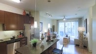 This single-floor space showcases a thoughtfully designed open-concept layout that seamlessly connects the kitchen and living areas. The kitchen is a standout feature, boasting rich dark cabinetry, granite countertops, and stainless steel appliances, including a French door refrigerator. A large center island with bar seating serves as both a functional workspace and a social hub, illuminated by elegant pendant lighting that adds warmth and style. Track lighting above provides additional flexibility for the space. The living area flows naturally from the kitchen, benefiting from abundant natural light streaming through large sliding glass doors that lead to an outdoor area, offering views of the surrounding greenery. A ceiling fan keeps the space comfortable year-round. The combination of polished flooring in the kitchen and plush carpeting in the living zone creates a clear yet cohesive transition between the two areas. Neutral wall tones throughout enhance the bright, welcoming ambiance, while thoughtful decorative touches — including ceramic accents and curated furnishings — contribute to a refined, contemporary aesthetic. The overall design balances practicality with modern style, making this an inviting and highly functional living environment.