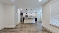 This two-story home showcases a contemporary design with clean lines and a neutral color palette throughout. The main level centers around an open-concept kitchen and living area, unified by durable wood-look flooring. The kitchen is well-equipped with white upper cabinetry, contrasting navy lower cabinets, a subway tile backsplash, stainless steel appliances, and a central island topped with granite countertops. Black industrial-style pendant lights hang above the island, adding a modern accent. Recessed lighting illuminates the space, which connects seamlessly to an open living area with windows dressed in white blinds that allow natural light to filter in.

The upper level features carpeted bedrooms, including a generously sized primary suite with two walk-in closets offering extensive built-in shelving and hanging rods. The primary bathroom is finished with wood-look flooring, a dark vanity with granite countertops, a backlit mirror, matte black fixtures with gold hardware accents, and a soaking tub. A secondary bathroom mirrors this refined aesthetic with a matching dark vanity, granite countertop, and framed mirror. A ceiling fan and staircase with a metal railing connect both levels cohesively.