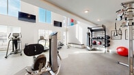 This well-appointed, two-story fitness center offers a comprehensive and thoughtfully designed workout environment across two connected levels. The main floor commands attention with soaring double-height ceilings and an abundance of natural light streaming through large clerestory windows, creating a bright and energizing atmosphere. This level features a versatile functional training zone equipped with a heavy bag, kettlebell rack, medicine balls, and a wall-mounted multi-purpose rig. Strength machines and cardio equipment, including treadmills and rowing machines, are strategically arranged throughout, while wall-mounted flat-screen televisions provide entertainment. Exposed light brick accent walls and a mix of carpet and hard flooring add warmth and texture to the crisp white interior. A staircase with sleek metal railings connects to the upper level, which houses a dedicated cardio area outfitted with air bikes, a stair climber, treadmills, and a full rack of dumbbells set against floor-to-ceiling mirrors that enhance the sense of space. A separate studio room on this floor features beautiful light wood plank flooring, a ceiling fan, decorative wall art, and a mounted television set against a striking reclaimed wood accent wall — ideal for yoga, stretching, or group fitness classes.