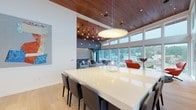 This single-floor space showcases a seamlessly integrated open-plan layout defined by striking architectural details and an abundance of natural light. Throughout, warm wood-paneled ceilings contrast elegantly with crisp white walls and light hardwood flooring, creating a refined yet welcoming atmosphere.

The expansive living area is anchored by floor-to-ceiling windows that span nearly the entire perimeter, framing sweeping panoramic views of the surrounding treeline and cityscape. A generous seating arrangement of neutral-toned sofas and accent chairs surrounds a marble-topped coffee table, while Edison-style pendant lights hang from the wood ceiling overhead.

Adjacent to the living area, the dining space features a large, glossy white table with seating for numerous guests, illuminated by a sculptural pendant fixture. Doors from this area open directly onto an outdoor terrace, furnished for alfresco dining and further extending the indoor-outdoor connection.

A dedicated media lounge offers a more intimate setting, complete with a wall-mounted large-format display, upholstered seating, a textured accent wall with integrated mood lighting, and the same warm wood ceiling detail.

The kitchenette area is equipped with stainless steel appliances, a deep undermount sink, white quartz countertops, and dark floating shelving for organized display and storage. Together, these spaces form a cohesive, contemporary environment designed for both comfort and functionality.