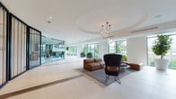 Located at 1057–1109 NW Seventh St in Miami, FL, this contemporary amenity floor presents a thoughtfully designed collection of communal spaces that blend modern architecture with refined interior details. The centerpiece is a sweeping lobby lounge characterized by large-format white tile flooring, a distinctive recessed circular ceiling detail, and globe pendant chandeliers that anchor the seating area. Expansive floor-to-ceiling windows line the perimeter, flooding the space with natural light and offering lush green outdoor views. Comfortable leather seating and layered area rugs create welcoming conversation areas throughout. A glass-partitioned wall with vertical wood slat accents elegantly divides the lounge from adjacent spaces, adding warmth and visual interest. The fully appointed kitchen features a substantial concrete-topped island with bar seating, marble-look backsplash, wood slatted cabinetry, stainless steel appliances, and artisan woven pendant lights. A casual dining and café nook showcases bold teal accent walls, built-in banquette seating, and tropical-print furnishings. The fitness center is visible through glass walls and offers professional-grade equipment. Light oak wood flooring, a dedicated reception desk area, and carefully curated artwork complete the polished, resort-inspired aesthetic of this single-floor amenity space.