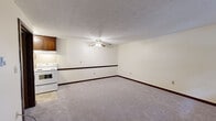 Located at 59–61 Meadow Brook Rd in Ellington, Connecticut, this single-floor unit offers a straightforward and functional layout with several notable design elements throughout. The living area is a generous, open space finished with neutral wall-to-wall carpeting and a ceiling fan with light fixture overhead, lending a comfortable, airy feel. A standout feature is the full-length exposed brick accent wall, which adds warmth and character to the space. Large sliding glass doors with vertical blinds allow natural light to fill the room while providing direct access to the outdoors and views of the surrounding greenery. The kitchen is efficiently arranged with rich, dark wood cabinetry, a white electric range, a full-size refrigerator, a stainless steel sink, and tile flooring — offering practical utility in a compact footprint. The bedroom is quietly carpeted and features warm-toned wood trim, with access to a dedicated closet providing ample storage. The bathroom is classically appointed with light blue tile wainscoting, a white vanity with mirrored medicine cabinet, a combination tub and shower, and bright overhead lighting. Exposed brick detailing carries into the bathroom as well, maintaining a cohesive design thread throughout the unit.