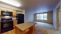 This single-floor unit offers a thoughtfully designed open-concept layout that seamlessly connects the kitchen and living area. The kitchen is fully equipped with warm honey-toned wood cabinetry, dark countertops, and a complete suite of black appliances including a range, microwave, dishwasher, and refrigerator. A central kitchen island provides additional prep space and doubles as a casual dining area. Light hardwood flooring runs throughout the kitchen, transitioning to soft, neutral carpeting in the spacious living area. Large windows dressed with vertical blinds fill the living room with natural light and offer views of the surrounding exterior. The bedroom is a comfortable, carpeted retreat with a dedicated closet featuring built-in shelving and direct access to the full bathroom. The bathroom is finished with tile flooring, a wooden vanity with an integrated sink, a large mirror, and ample towel storage. The overall ambiance of the unit is clean, functional, and welcoming, with a neutral warm color palette and practical finishes throughout that make it a versatile and move-in ready space.
