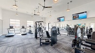 Located at 9944 Highway 6 in Missouri City, TX, this fitness center offers a generously sized, single-floor exercise room designed to support a comprehensive workout experience. The space features soaring ceilings that create an open, airy atmosphere, complemented by multiple large windows that flood the interior with natural light and provide outdoor views of the surrounding area. Stylish pendant lights and ceiling fans are suspended overhead, enhancing both comfort and visual appeal.

The exercise room is finished with durable, patterned carpet tiles and white shiplap-style walls that lend a clean, modern aesthetic. Full-length mirrors line one wall, amplifying the sense of space and allowing users to monitor their form during workouts.

The facility is outfitted with a diverse array of equipment, including Instinct-brand strength machines, a cable crossover system, adjustable benches, and a full dumbbell rack with medicine balls for free-weight training. Cardio options are equally well-represented, with treadmills, ellipticals, a stair climber, and stationary bikes positioned throughout the room. Plyo boxes add functional training versatility. Multiple wall-mounted flat-screen televisions are positioned for easy viewing from across the floor, making workouts more engaging. The overall ambiance is bright, modern, and motivating.