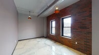 Located at 204 E Grand Ave in downtown Eau Claire, Wisconsin, this single-floor urban loft-style unit showcases a striking blend of industrial and modern design elements throughout. The main living area is a standout, featuring a rich exposed red brick accent wall punctuated by black-framed windows that frame views of the surrounding cityscape, filling the space with natural light. Polished marble-effect epoxy floors run seamlessly throughout, adding a sleek, contemporary contrast to the raw industrial aesthetic. Exposed cylindrical ductwork traces the ceiling, complemented by pendant lights with warm copper-toned shades that provide ambient illumination. The kitchen is well-appointed with dark espresso cabinetry, stainless steel appliances including a range, over-the-range microwave, and refrigerator, along with a granite-topped island featuring a deep double stainless steel sink. The bathroom includes granite countertops, a full tub-shower combination, and continues the cohesive flooring found throughout the unit. Additional rooms offer flexible space, with tall ceilings, neutral gray walls, and dark-framed interior doors maintaining the industrial-modern character. A utility closet houses the water heater, conveniently tucked away. The overall ambiance is bold, contemporary, and well-suited for urban living.