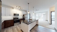 This single-floor unit offers a well-designed, open-concept layout that seamlessly connects the kitchen, dining, and living areas. The kitchen is a standout feature, equipped with stainless steel appliances including a refrigerator, electric range, and built-in microwave. Two-tone cabinetry pairs rich dark-wood lower cabinets with crisp white upper cabinets, complemented by a classic white subway tile backsplash and dark granite countertops. A central island with an undermount sink, brushed gold faucet, and pendant lighting above serves as both a functional workspace and a natural gathering point. Light wood-style flooring runs throughout the kitchen and main living areas, while plush carpeting covers the living room and bedrooms.

The spacious living area is bathed in natural light from large sliding glass doors that open onto a private balcony overlooking the surrounding neighborhood. Two bedrooms offer comfortable proportions, each with generous windows. The unit includes two bathrooms: one with a tiled tub-shower combination and one with a glass-enclosed shower, both featuring granite-topped vanities with dark wood cabinetry. A dedicated laundry room houses a full-size top-load washer and dryer with wire shelving above for additional storage.