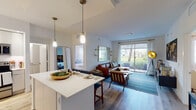 This single-floor residence showcases a thoughtfully designed open-concept layout that seamlessly connects the kitchen, dining, and living areas. The kitchen is outfitted with flat-front cabinetry in white and light gray, quartz countertops, stainless steel appliances, under-cabinet lighting, and a central island with bar seating illuminated by brass pendant lights. Light wood-tone flooring flows consistently throughout the entire unit, enhancing the cohesive, airy atmosphere.

The living room is anchored by a sliding glass door that opens to a private patio, framing views of green landscaping and palm trees. Natural light fills the space generously, complementing the neutral white walls and modern furnishings.

The primary bedroom features a ceiling fan, a walk-in closet, and a dedicated en-suite bathroom with large-format tile flooring, a walk-in glass shower with gray subway tile surround, granite vanity countertops, and recessed lighting. A second bedroom, equally bright and well-proportioned, also benefits from natural light through a large window and includes a ceiling fan. Both bedrooms share the same warm wood-tone flooring found throughout the residence, contributing to a consistent and polished aesthetic.
