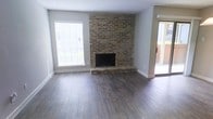 This single-floor space combines classic and contemporary design elements to create a comfortable and welcoming environment. The living area serves as the heart of the space, anchored by a full-height brick fireplace that commands attention as a bold architectural focal point. The rich, textured brickwork adds warmth and character, complementing the smooth grey walls and crisp white trim throughout.

Underfoot, wide-plank wood-look flooring in a rich grey-brown tone runs throughout the space, providing both durability and a modern aesthetic. The flooring ties the room together and enhances the overall sense of warmth.

Natural light plays an important role in the ambiance of the space. A large window fitted with white horizontal blinds allows generous daylight to fill the room, brightening the interior while offering privacy as needed. A sliding glass door provides a seamless connection to an outdoor area, extending the living space beyond its interior boundaries and inviting fresh air and additional light.

The open layout of the living area offers flexibility for a variety of furniture arrangements, making it adaptable to different lifestyle needs. The textured ceiling adds subtle visual interest overhead, while the clean lines and neutral palette of the space create a versatile backdrop that suits a range of interior design styles.