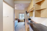 This single-floor studio unit at 1505 N Fourth St in San Jose, California, offers a compact and efficiently organized layout designed to maximize functionality. The main living area combines sleeping and living space, featuring carpeted flooring, a ceiling fan, and a window that allows natural light to filter in while providing views of surrounding greenery. The sleeping area is furnished with a bed, a small armchair, and side tables, with wall-mounted sconce lighting adding a warm ambiance. Decorative framed artwork on the walls contributes a welcoming touch.

A built-in kitchenette lines one wall, outfitted with light wood cabinetry—both upper and lower—providing ample storage. The countertop area includes a stainless steel sink, a compact cooktop, a microwave, and a mini-refrigerator. A wall-mounted television and a dedicated desk area with drawer storage make the space practical for everyday use or extended stays.

The bathroom features tile flooring in a natural stone finish, a wall-mounted sink, a toilet, a mirrored medicine cabinet, and a full bathtub with a shower, complete with grab bars for added accessibility. The overall design is clean, neutral, and functional, well-suited for single-occupancy use.