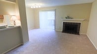 This single-floor space offers a clean, comfortable layout designed for easy living. The living area serves as the heart of the home, featuring soft wall-to-wall carpeting in a neutral tone that adds warmth underfoot. A standout feature of the room is the classic fireplace, framed by a dark tile surround and topped with a crisp white mantel — an inviting focal point ideal for relaxing evenings. Natural light flows generously into the space through a large sliding glass door dressed with vertical blinds, offering both privacy and easy access to the outdoors. The walls are finished in a soft, muted tone that complements the overall light and airy atmosphere of the room. A multi-bulb ceiling light fixture provides ambient illumination, enhancing the welcoming feel of the space. The textured ceiling adds subtle architectural character. The open layout of the living area allows for flexible furniture arrangements, making it adaptable to a variety of lifestyle needs. Overall, this space presents a practical yet comfortable environment with key features — including the fireplace, natural light, and neutral finishes — that combine to create a relaxed and functional living experience.