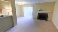 This single-floor space offers a clean, comfortable layout designed for easy living. The living area serves as the heart of the home, featuring soft wall-to-wall carpeting in a neutral tone that adds warmth underfoot. A standout feature of the room is the classic fireplace, framed by a dark tile surround and topped with a crisp white mantel — an inviting focal point ideal for relaxing evenings. Natural light flows generously into the space through a large sliding glass door dressed with vertical blinds, offering both privacy and easy access to the outdoors. The walls are finished in a soft, muted tone that complements the overall light and airy atmosphere of the room. A multi-bulb ceiling light fixture provides ambient illumination, enhancing the welcoming feel of the space. The textured ceiling adds subtle architectural character. The open layout of the living area allows for flexible furniture arrangements, making it adaptable to a variety of lifestyle needs. Overall, this space presents a practical yet comfortable environment with key features — including the fireplace, natural light, and neutral finishes — that combine to create a relaxed and functional living experience.