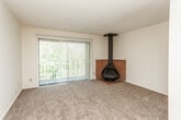 This single-story residence offers a well-defined layout connecting several functional living areas. The living room serves as the central gathering space, finished with plush carpet and anchored by a striking freestanding wood-burning fireplace with a bold black flue. A full-width sliding glass door opens directly onto a private balcony enclosed by a wrought iron railing, framing views of mature surrounding trees and greenery. Natural light flows generously into this space through the glass door and its accompanying vertical blinds.

Adjacent to the living room, the kitchen is outfitted with warm oak cabinetry, laminate countertops, a double-basin sink, and white appliances including a range and dishwasher. Wood-look flooring runs through the kitchen and entry areas, providing a practical contrast to the carpeted living and bedroom spaces.

The bedrooms are comfortably sized and naturally lit through windows fitted with blinds. Each bedroom includes closet storage with rod and shelf systems. The bathrooms feature white tile tub-and-shower combinations with accent tile detailing, wood-toned vanity cabinetry, and wood-look flooring. A secondary bath offers a dedicated shower stall. Textured ceilings are present throughout the unit.