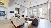 Located along Essex Lane in Houston's upscale 77027 corridor, this single-floor community amenity and leasing space is thoughtfully designed to offer both function and comfort. The entrance opens into a welcoming lobby featuring warm hardwood-style flooring, bold globe chandeliers, and a seating area with crisp white armchairs accented by royal blue pillows, set atop a striking blue-and-white abstract area rug. Adjacent to the lobby, the leasing office is finished with teal accent walls, open display shelving, elegant pendant lighting, and a sleek front desk with guest seating — creating a professional yet inviting atmosphere. The expansive lounge area impresses with soaring ceilings, a floor-to-ceiling white brick fireplace with a wall-mounted television above, and large multi-pane windows that flood the space with natural light and offer views of the surrounding urban landscape. A sputnik-style gold chandelier crowns the seating arrangement, which includes a sectional sofa, accent chairs, and a geometric coffee table. A dedicated dining area with a full table and chairs sits beside the windows. The kitchen features warm wood cabinetry, a textured tile backsplash, stainless steel appliances including a French door refrigerator, and a built-in workstation with a printer — ideal for residents and staff alike.