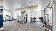 Located at 2005 S 300 W in Salt Lake City, Utah, this fitness center offers a thoughtfully designed exercise space across a single floor. The room is finished with neutral-toned carpet tiles that provide both comfort and durability underfoot, while crisp white walls and a textured ceiling contribute to a clean, modern aesthetic.

Natural light streams in through a bank of large windows fitted with horizontal blinds, offering views of the surrounding area and creating an open, airy atmosphere. Recessed lighting throughout ensures the space remains well-lit at all times of day.

The exercise room is comprehensively equipped to support a full-body workout routine. Cardio equipment includes treadmills, an elliptical, a stationary bike, and a rowing machine, all positioned to take advantage of the natural light from the windows. A multi-station cable machine and various strength training apparatus provide ample options for resistance training. A dedicated free weights and functional training area features a dumbbell rack, exercise mat flooring, stability balls, and TRX suspension straps. Wall-mounted televisions are positioned for easy viewing during workouts.

A set of double doors leads to an adjacent utility area, and hand sanitizer stations and cleaning supplies are conveniently mounted near the entrance, emphasizing a clean and hygienic environment for all users.