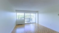This single-floor space offers a clean, minimalist aesthetic defined by white walls and warm parquet wood flooring laid in a classic block pattern throughout. The living area is the focal point of the space, benefiting from an abundance of natural light that streams in through a wide floor-to-ceiling sliding glass door system spanning much of the exterior wall. This generous glazing not only floods the interior with daylight but also provides direct access to a private balcony, which overlooks a lush canopy of mature trees — a notably tranquil and green outlook. The elevated position of the unit enhances the sense of openness, offering unobstructed views of the surrounding treetops and sky. Inside, the living area is spacious and versatile, with smooth white ceilings and walls that create a neutral backdrop adaptable to a variety of furnishing styles. A baseboard heating unit is positioned beneath the windows, ensuring comfort year-round. The overall ambiance of the space is bright, airy, and calm, with the natural surroundings visible through the expansive windows contributing to a serene atmosphere well-suited for comfortable everyday living.