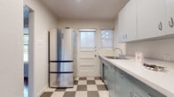 This single-floor home presents a thoughtfully arranged layout with a blend of classic and retro design elements throughout. The living areas are defined by rich, dark hardwood floors that run continuously, reflecting natural light and lending warmth to the open, neutral-toned spaces. Exposed beam detailing accents the ceiling in the main living area, adding architectural character, while large windows fitted with roller shades allow for generous natural light and outdoor views.

The kitchen carries a distinctly retro aesthetic, featuring powder-blue metal cabinetry paired with white countertops and a classic checkerboard vinyl floor in grey and cream tones. A stainless steel refrigerator and a gas range complete the functional cooking space, while a door with a windowed panel provides direct exterior access and brings additional natural light into the room.

The bathroom is a standout feature, showcasing floor-to-ceiling turquoise ceramic tile surrounding a full bathtub, complemented by a small window above — a bold, vintage-inspired design choice that gives the room a distinctive personality.

Additional rooms include well-proportioned bedrooms with hardwood flooring and large windows offering outdoor views, along with built-in shelving visible in the doll's-eye floorplan view, providing practical storage solutions throughout the home.