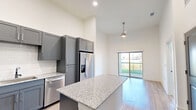 Located in Denton, TX, this well-appointed single-story residence offers a thoughtfully designed open-concept layout. The combined kitchen and living area serves as the heart of the home, highlighted by soaring ceilings that create an airy, expansive feel. The kitchen is outfitted with sleek gray shaker cabinetry, granite countertops, a subway tile backsplash, a central island with additional counter space, and a full suite of stainless steel appliances. Light wood-look flooring flows throughout the main living space, while a ceiling fan and a sliding glass door with outdoor views complete the living area. The spacious bedroom features plush carpet, dramatic ceiling height, and large windows that flood the room with natural light. It connects directly to the full bathroom, which includes a dark navy vanity with granite countertops, a framed mirror, and a separate walk-in shower. Four closets throughout the unit provide generous storage. A dedicated laundry area adds everyday convenience. The consistent use of neutral tones, modern fixtures, and high-end finishes gives the space a clean, contemporary ambiance throughout.