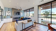 Located at 70 N Winchester Blvd in Santa Clara, California, this single-floor space presents a thoughtfully designed contemporary interior that blends comfort with functionality. The layout flows seamlessly from a welcoming lounge area into multiple distinct seating and dining zones, all unified by warm wood-look flooring and crisp white walls.

The lounge area serves as the centerpiece of the space, anchored by a striking floor-to-ceiling herringbone-tiled fireplace wall with a wall-mounted television above a sleek linear hearth. Comfortable seating, including a light gray sofa and navy blue leather armchairs, creates an inviting atmosphere, while pendant lighting and recessed fixtures provide layered illumination throughout.

Large black-framed windows line the perimeter, flooding the interior with generous natural light and offering views of the surrounding outdoor landscaping, including mature trees and a courtyard area. A glass-panel opening connects the interior to the outdoors, enhancing the sense of openness.

The kitchen features white cabinetry, dark countertops, a textured tile backsplash, and stainless steel appliances including a refrigerator, range, and range hood. Adjacent dining areas offer both casual round marble-topped tables and a more formal rectangular conference-style table, accommodating a variety of uses throughout the day.