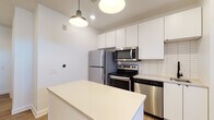 Located at 500 W Broad St in Columbus, Ohio, this contemporary single-floor unit offers a thoughtfully designed layout with modern finishes throughout. The kitchen is a standout feature, showcasing crisp white flat-front cabinetry extending to the ceiling for ample storage, a full suite of stainless steel appliances including a refrigerator, electric range, over-the-range microwave, and dishwasher, complemented by a white subway tile backsplash and light quartz countertops. A central island provides additional prep space and casual seating. Stylish pendant lighting adds a modern touch overhead. The open living area is finished with warm-toned luxury vinyl plank flooring and bright white walls, creating a clean, airy atmosphere. Recessed lighting throughout ensures consistent illumination. French doors with transom windows invite abundant natural light and provide access to a private balcony with views of the surrounding Columbus streetscape. The bathroom is finished with a sleek white vanity, matte black hardware accents, a large mirror, and distinctive dark hexagonal mosaic tile flooring, paired with a tiled walk-in shower. The overall design aesthetic is modern and cohesive, blending functionality with contemporary style in a prime Columbus location.